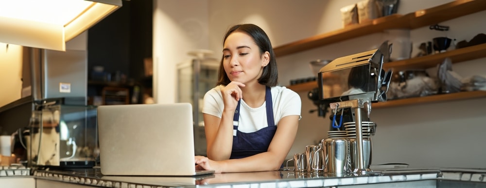 Portrait of smiling korean woman, barista in coffee shop, standing at counter with laptop, smiling and looking confident, self-employed female entrepreneur in her own coffee shop.